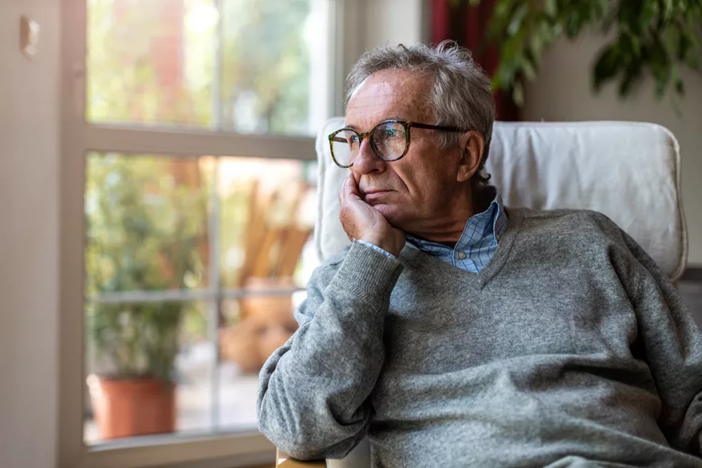 An older adult man looking somber, sitting in his living room, struggling with the mental health effects of untreated hearing loss